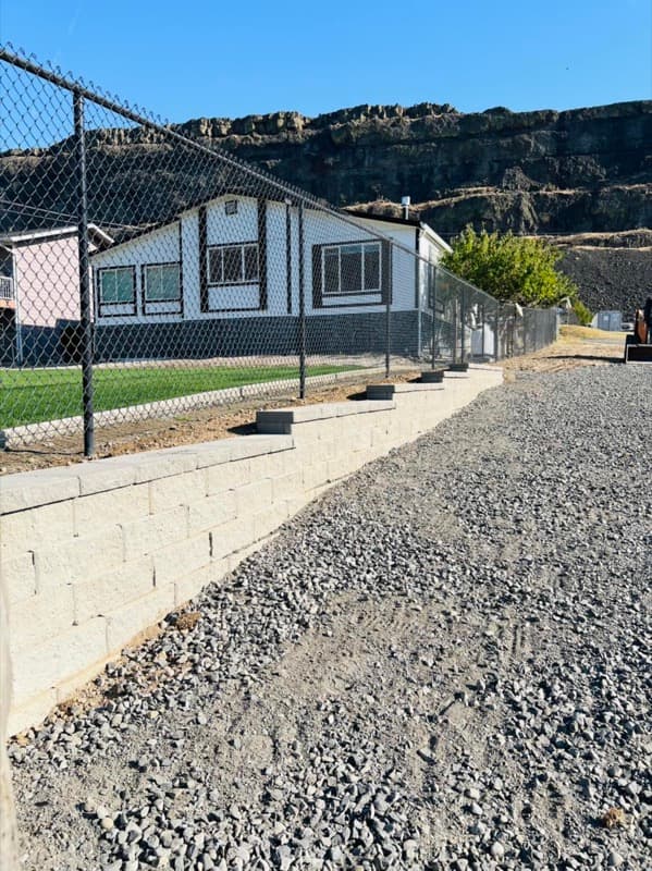 Muro de contención de bloques junto a un camino de grava bajo una cerca de malla ciclónica