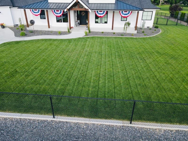 Freshly mowed striped lawn in front of a white farmhouse with a black fence